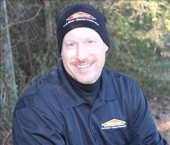 man outside wearing a black jacket and beanie smiling, there are leaves and trees behind him