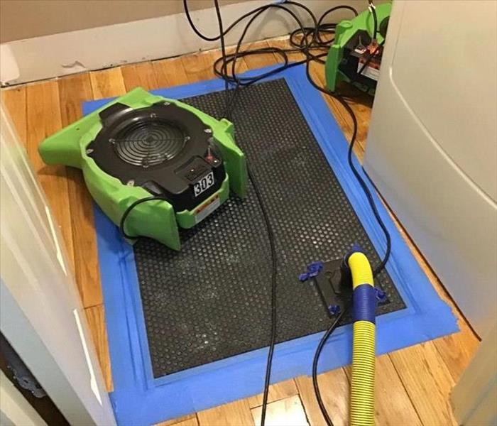 A wood floor with blue tape holding down a black pad with yellow tubes coming out of it. There is a green air mover at the to